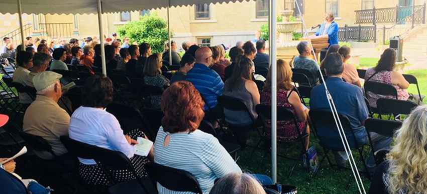 Group of people under a tent outside listening to a speaker at a community event