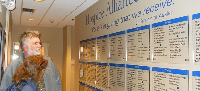 A man and woman standing next to the Hospice Alliance donor wall showing different levels of donation